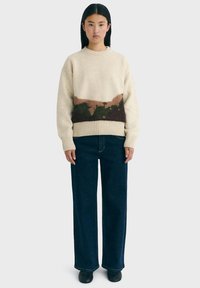 Woman standing wearing a cream sweater with brown and green pattern near the waist, dark wide-leg jeans, and black shoes on a plain background.