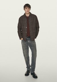 Brunternet ternet jakke over en bordeaux sweater, parret med lyse slidte jeans og sorte læderstøvler. Model står imod en ensfarvet baggrund.