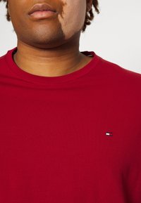 Red cotton t-shirt featuring a round neck and a small logo on the chest, displaying a blue, white, and red color scheme. Smooth texture.