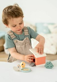 Casă de joacă din lemn, portocalie, cu un acoperiș alb; include o rampă gri și figurine de animale (pasăre și leu), plus accesorii sub formă de bol și tufiș.