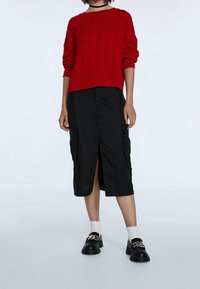 Pull en maille rouge avec des poignets côtelés, associé à une jupe cargo noire mi-longue avec des poches latérales et une fente à l'avant. Mocassins noirs épais.