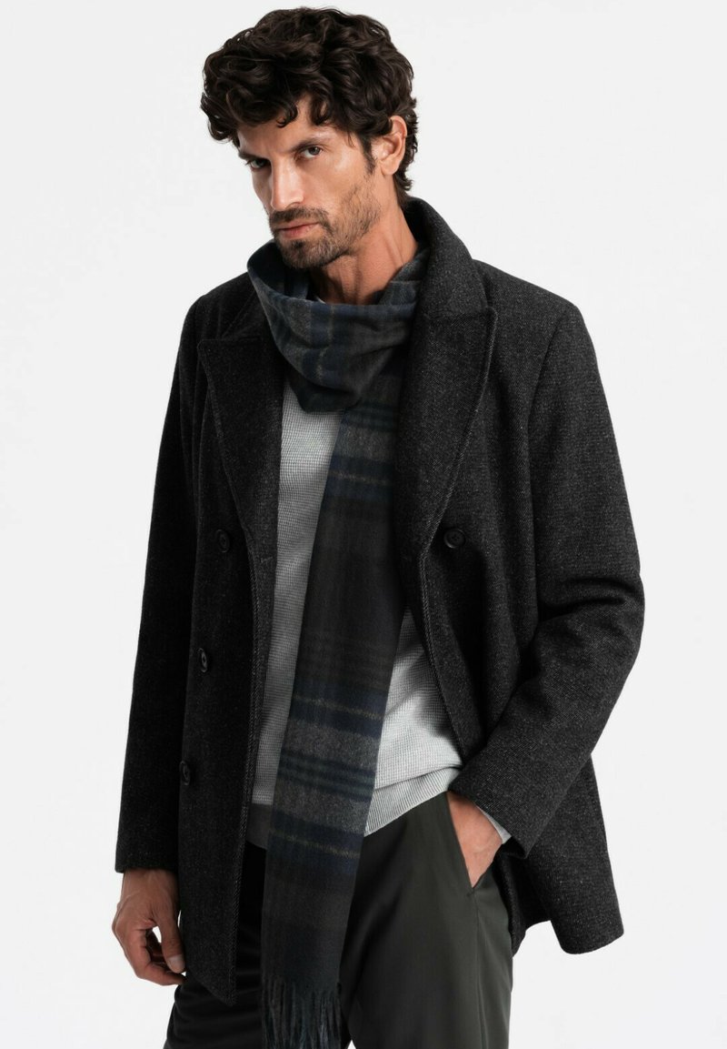 Grå uldfrakke med dobbeltradet design, sorte knapper og en tekstureret overflade, parret med et mørkt rutet tørklæde. En lys sweater underneath.