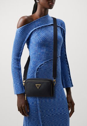 Mujer con un vestido azul de punto acanalado y hombros descubiertos, llevando un bolso cruzado negro de Guess con herrajes dorados y logo en la parte frontal.
