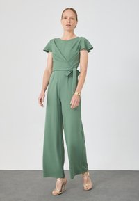 Grön jumpsuit med korta flamboyant ärmar, omlottdesign och sidbanddetalj. Slät textur och vida ben, parat med guldklackar med remmar.