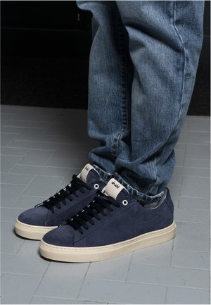 Person wearing blue suede sneakers with black laces and white soles, paired with light blue denim jeans standing on gray tiled floor.