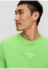 Grünes Rundhals-T-Shirt aus weicher Baumwolle mit lockerer Passform. Mit gesticktem Logo in hellem Creme auf der Brust. Sanfte Textur.