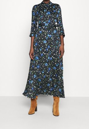 Femme portant une robe maxi à manches longues noire à fleurs bleues et blanches, assortie à des bottes à talons en daim beige.
