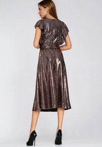 Kobieta w błyszczącej, metalicznej sukience midi z krótkim rękawem i falbanami, stojąca tyłem, w czarnych szpilkach.
