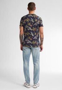 N�ámořnické tričko s tropickým vzorem palmy, kombinované se světle modrými džíny a bílými teniskami. Tetovaná ramena viditelná zezadu.