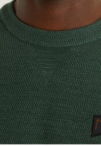 Grüner strukturierter Pullover mit Rundhalsausschnitt, geripptem Kragen und einem kleinen schwarzen Logo-Tag mit orangefarbenen Akzenten.