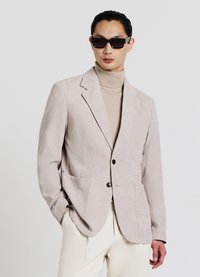 Beiger Cordblazer mit zwei Vordertaschen, Ein-Knopf-Verschluss, Revers mit Kerbung und strukturierter Stoff. Über einem beigen Rollkragenpullover getragen.