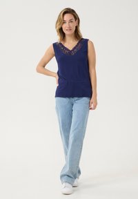 Top senza maniche di colore blu navy con dettagli in pizzo al collo a V, abbinato a jeans azzurri e scarpe da ginnastica bianche, in piedi di fronte a uno sfondo neutro.