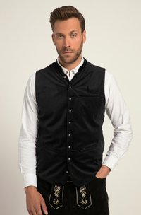 Zwarte suède vest met hoge kraag, voor knopen en twee zakken, gedragen over een wit shirt, gecombineerd met geborduurde zwarte broeken.