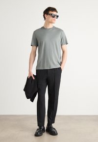 Tricou gri cu mânecă scurtă, pantaloni negri croiți, mocasini negri, ținând o jachetă neagră, purtând ochelari de soare închiși la culoare. Material neted, design simplu.
