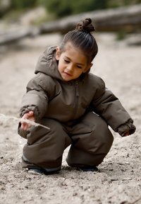 Bruine geïsoleerde skipak met capuchon, elastische manchetten en zijzakken. Kind hurkt op zand en houdt een slanke stok vast.