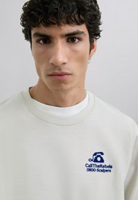 Sweatshirt bej deschis cu guler rotund, având un grafic imprimat albastru cu un telefon și textul "CallTheRebels 0800-Scalpers."