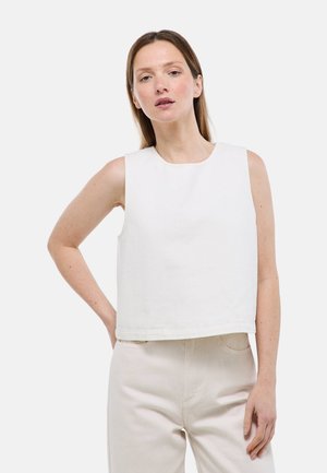 Femme à la peau claire et aux cheveux bruns raides portant un haut blanc sans manches et un pantalon beige clair, debout avec la main sur la hanche devant un fond uni.