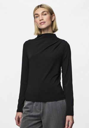 Long-sleeve black top made of smooth fabric with a high collar and draped detail on the shoulder, paired with gray trousers.