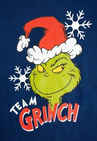Maglietta blu con un personaggio Grinch verde che indossa un cappello da Babbo Natale rosso e bianco, con il testo "TEAM GRINCH" e fiocchi di neve bianchi.