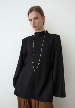 Kobieta o jasnych włosach, ubrana w luźną czarną bluzkę z długim rękawem i brązowe spodnie, z długim srebrnym naszyjnikiem na jednolitym tle.