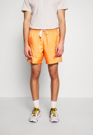 Homme portant un short Nike orange vif avec un cordon blanc, des chaussettes blanches montantes et des baskets blanches avec des accents jaunes et noirs, debout sur fond blanc.