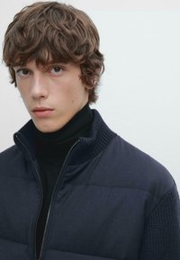 Mladý muž s vlnitými hnědými vlasy, oblečený v námořnickém prošívaném kabátu přes černý rolák, na jednolitém světle šedém pozadí.