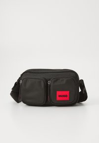 Schwarze Stoff-Crossbody-Tasche mit zwei Reißverschlusstaschen auf der Vorderseite, einem auffälligen roten Logopatch und einem verstellbaren Tragegurt. Glatte Textur, kompaktes Design.
