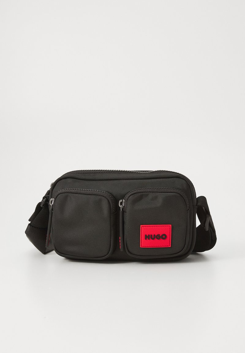 Svart stoff crossover-veske med to glidelåslommer foran, en fremtredende rød logo-lapp, og en justerbar stropp. Jevn tekstur, kompakt design.