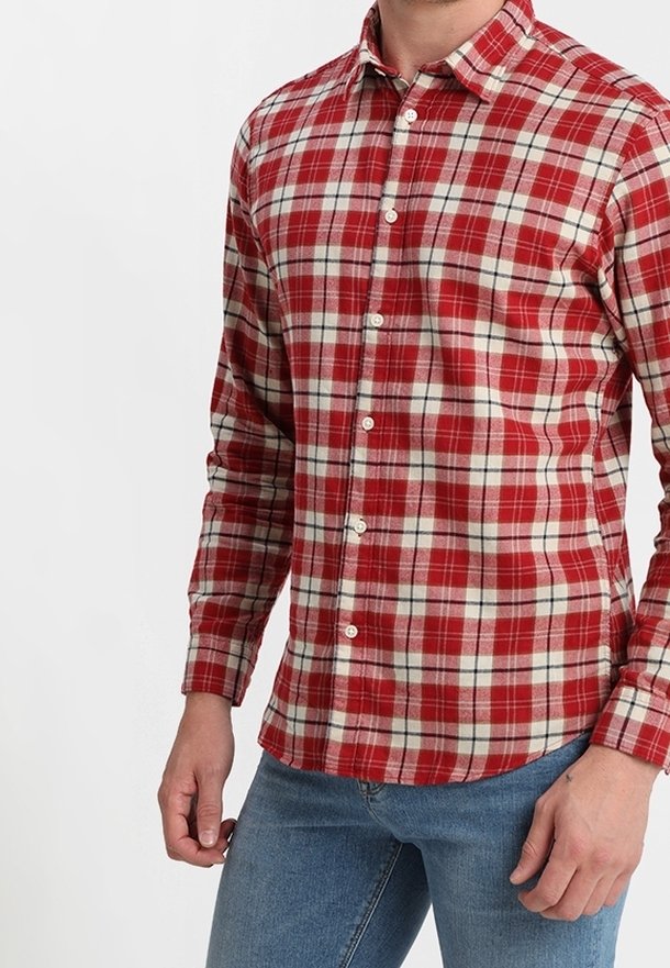 Homme portant une chemise à carreaux rouge et blanche à manches longues boutonnée, associée à un jean bleu, debout, les bras détendus sur les côtés.