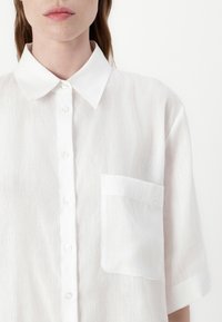 White, semi-sheer button-up shirt with a pointed collar, short sleeves, and a chest pocket. Subtle texture with a clean design.