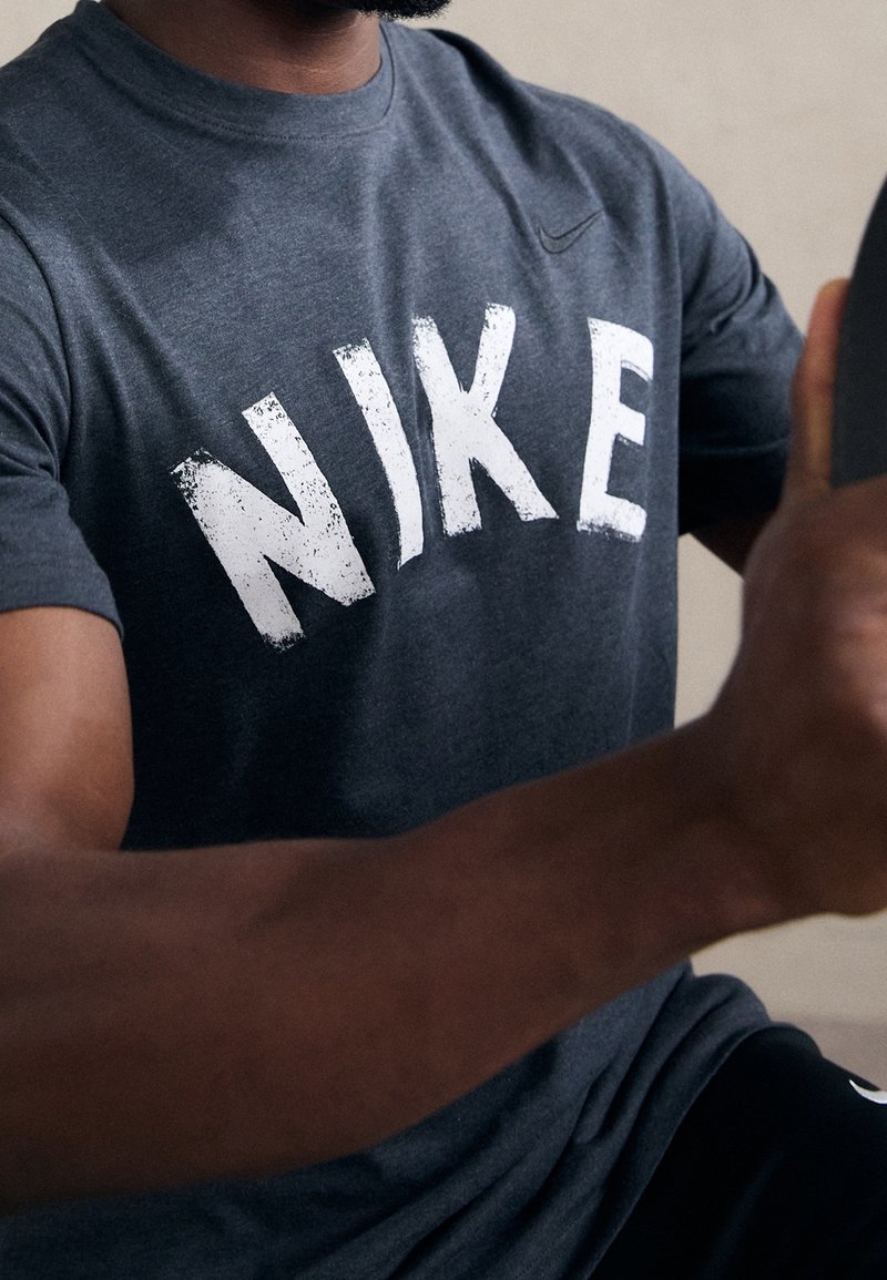 Šedé tričko s kulatým výstřihem, které na přední straně zdobí velké, opotřebované bílé logo "NIKE". Materiál vypadá měkký a má hladkou strukturu.