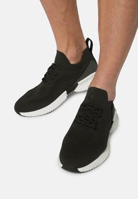 Svarta sportskor med ett stickat ovandel, texturerat mönster, vit sula och svarta accenter. Har snörning och ett dragöglan på hälen.