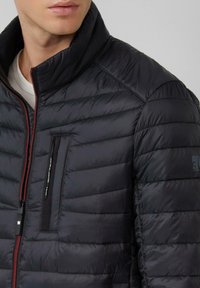 Zwarte gewatteerde jas met een hoge kraag, voorzien van horizontale stiknaden, een borstzak met rits en rode ritsaccenten. Glad, lichtgewicht materiaal.