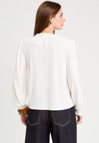 Blouse blanche à manches longues avec un col volanté et des manches rassemblées. Portée avec un pantalon en denim foncé, accentuée par un bracelet chunky.