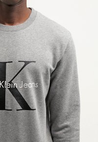 Sweat-shirt gris en mélange de coton avec un col rond. Présente un grand logo "K" noir et le texte "Klein Jeans" dans une police contrastante.