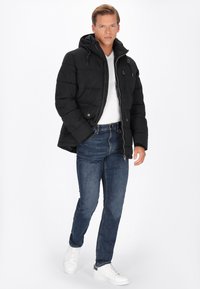 Geacă puffer neagră cu glugă, închidere cu fermoar și buzunare cu clapetă. Purtați peste un tricou alb, cu blugi albaștri și adidași albi.