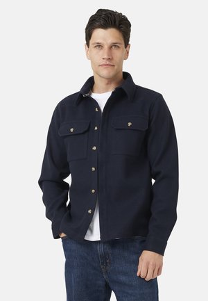 Man draagt een marineblauwe jas met knopen en twee borstzakken over een wit overhemd en blauwe spijkerbroek, staand met één hand in zijn zak.