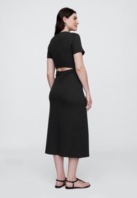 Črna midi obleka s kratkimi rokavi, izrezom na hrbtni strani in oprijetim krojem. Iz mehke, raztegljive tkanine. Poudarjena s športnimi sandali.