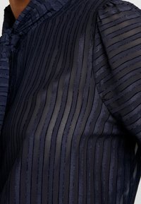 Námořnická modrá průsvitná blůza s vertikálními sametovými pruhy a zavázanou mašlí na výstřihu, nošená na horní části trupu osoby.