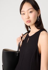 Abito nero senza maniche con scollatura a chiave; modella che tiene una borsa nera testurizzata con cuciture bianche e una forma strutturata.
