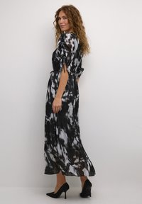 Zwart-wit tie-dye maxi jurk met geplooide textuur, taille met elastiek en puffed mouwen, gecombineerd met zwarte hoge hakken.