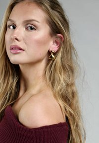 Guldörhängen med en abstrakt, flytande hjärtform. Modellen har l�ångt vågigt hår och bär en vinröd off-shoulder tröja.