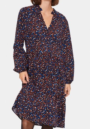 Femme portant une robe à manches longues et ample avec un motif tacheté multicolore en bleu, marron et blanc sur un fond sombre.