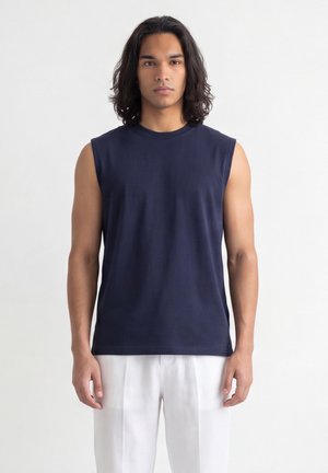 Giovane uomo con capelli scuri fino alle spalle, che indossa una canotta blu navy e pantaloni bianchi, in piedi su uno sfondo chiaro.