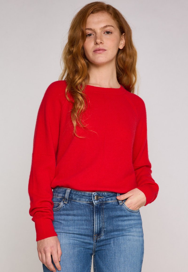 Maglione rosso lavorato a maglia con maniche lunghe e scollatura rotonda, abbinato a classici jeans in denim blu. Il maglione presenta una consistenza morbida.