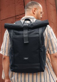 Man in een zwarte roll-top rugzak met een gesp, gestreept shirt met korte mouwen, staand tegen een roestkleurige muur.