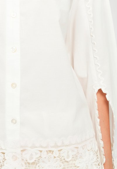 White buttoned garment with scalloped edges and floral lace trim at bottom, showing part of a forearm through side slit.