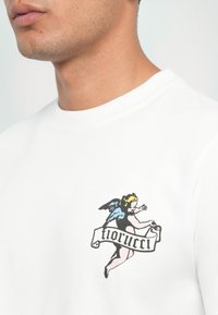 White cotton T-shirt featuring a colorful angel graphic with a banner reading "tiorucci" in bold text, showcasing a smooth texture.