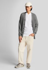 Šedý button-up kardigán, biele polo tričko, krémové nohavice, svetlo šedé tenisky s akcentmi. Oblečenie dopĺňa béžová čiapka.