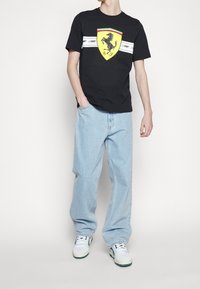 Svart bomullströja med en stor gul Ferrari-emblem, med vit och grön rand. Bärs med ljusblå lösa jeans och vita sneakers.